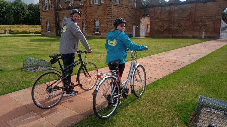 A man and girl wheeling bikes at Chatelherault Park, wearing Bikeability jackets.