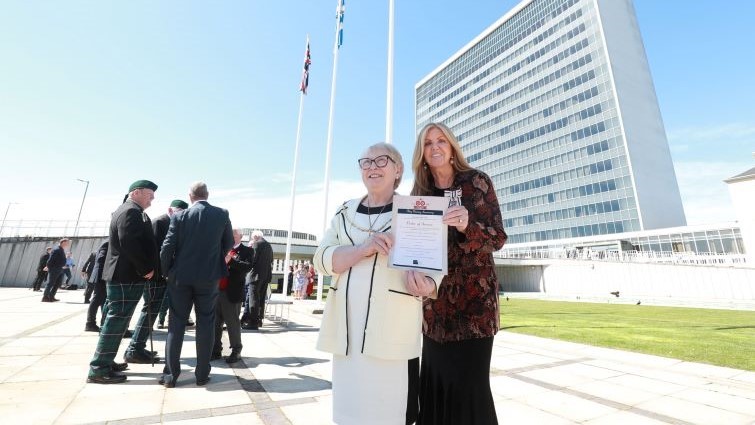 Provost Cooper and Lady Haughey stand together with the raised flag behind them and the council HQ building in the background.