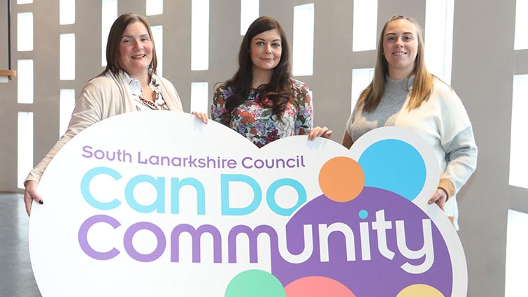 Three female members of the community engagement team stand side by side holding a large circular prop bearing the team logo 'can do community'.