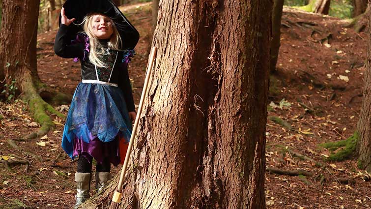 Alt text: This photo shows a little girl dressed as a witch, peeking out from behind the trees at Chatelherault.