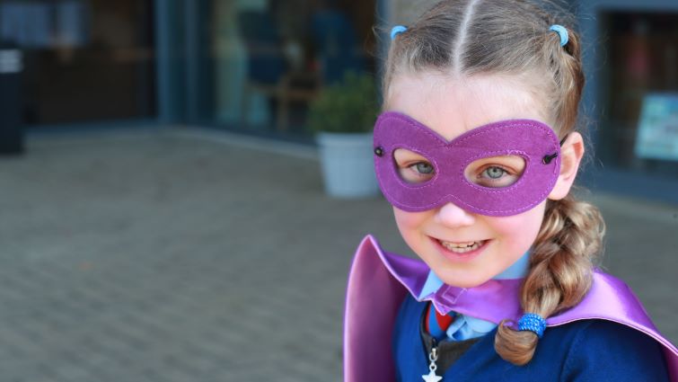 Primary one pupil Ellie is pictured outside school in her uniform wearing a purple mask and superhero cloak.