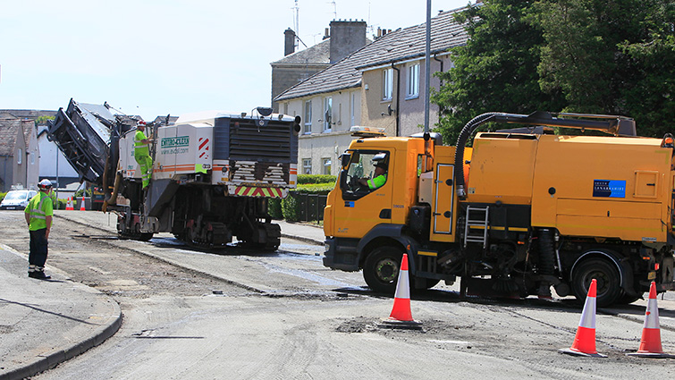 Two road resurfacing vehicles working on a coned off road