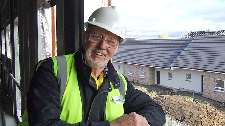 Councillor John Ross is leaning out of a window space on the upper floor of the care hub under construction in Blantyre. Behind him new council housing can be seen. He is wearing a white hard hat and yellow hi vis vest