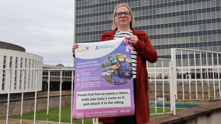 This image shows Councillor Lynsey Hamilton outside council HQ holding a banner highlighting Baby Loss Awareness Week and the opportunity to place a ribbon to remember babies gone too soon