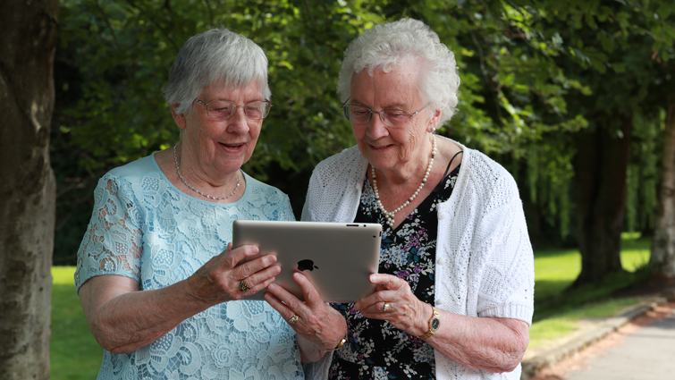 Two older women standing together and looking at an ipad.
