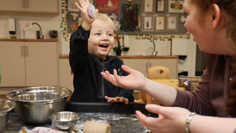 This image shows a young nursery pupil holding a piece of plasticine in the air as he hands it to a teacher in the foreground