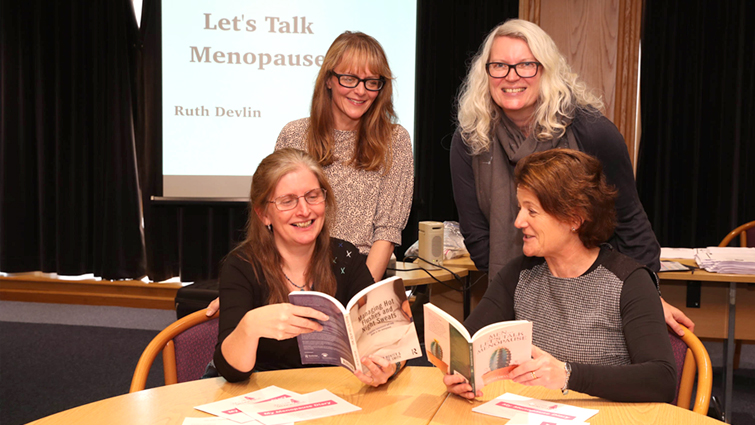 This photo shows a group of four female employees two seated and two standing behind looking through literature on the menopause and chatting to each other.