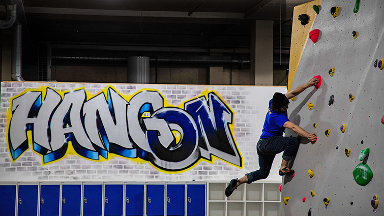 on the right of the image a man is hanging by his hands on a climbing wall while swinging his foot to reach a new hold. In the background a large grafitti style sign reads Hang On