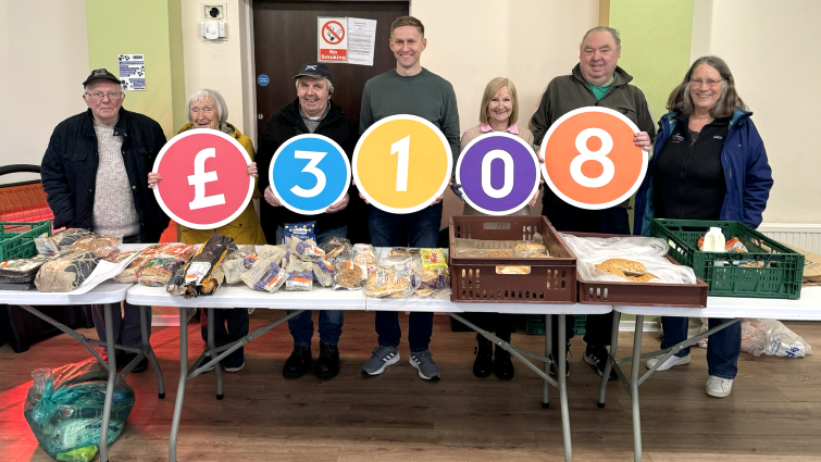 Seven members of the foodbank committee stand behind a table full of food. Five of them are holding colourful boards which together spell out £3108.