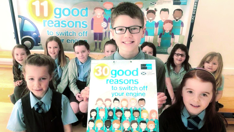 pupils in the Kirklandpark Primary School Eco Committee stand behind a poster that reads 30 reasons to switch off your engine, and has a graphic of a class of school pupils.