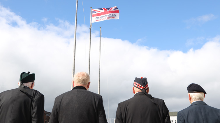 This image shows the backs of veterans looking on as the Armed Forces Day flag is raised on 25 June 2022
