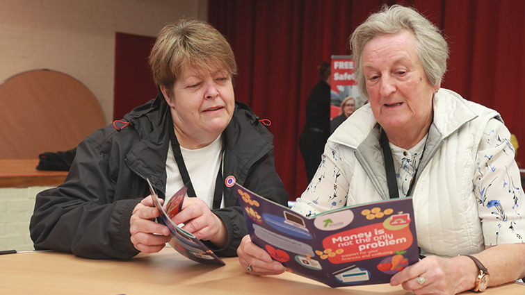 This image shows two people at a networking event in Ballerup Hall reading the Money is Not the Only Problem booklet
