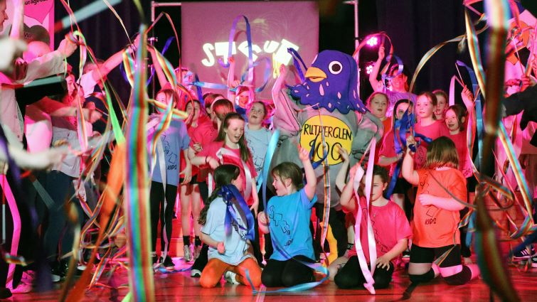 This is a group pic of the young people on stage at the end of the show. they are brightly dressed, waving ribbons and laughing. In the centre is Rutherglen 900 mascot, Dougie the pigeon.
