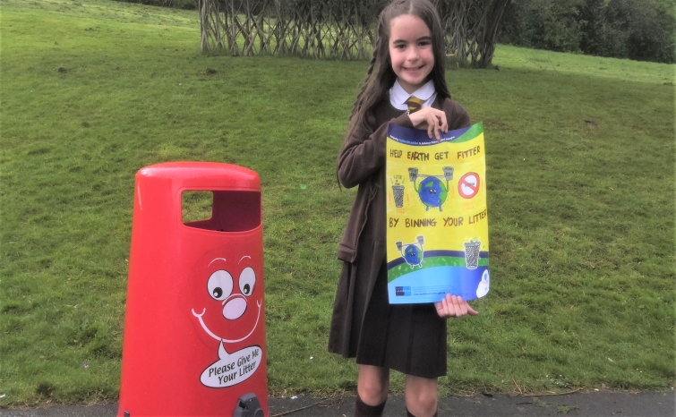 This photo shows competition winner, Sophia, standing in the school playground holding her winning poster in A3 size, She is standing next to one of the school's red litter bins.